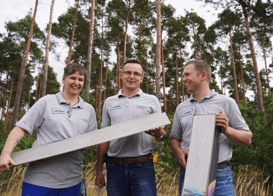 Drei CLASSEN Mitarbeiter in grauen Polo-Shirts mit Firmenlogo stehen lachend im Freien vor einem Wald und halten nachhaltige Bodenbeläge in den Händen. Das Foto symbolisiert umweltfreundliche Produktion, Teamarbeit und die Verbindung von Natur und moderner Fertigung
