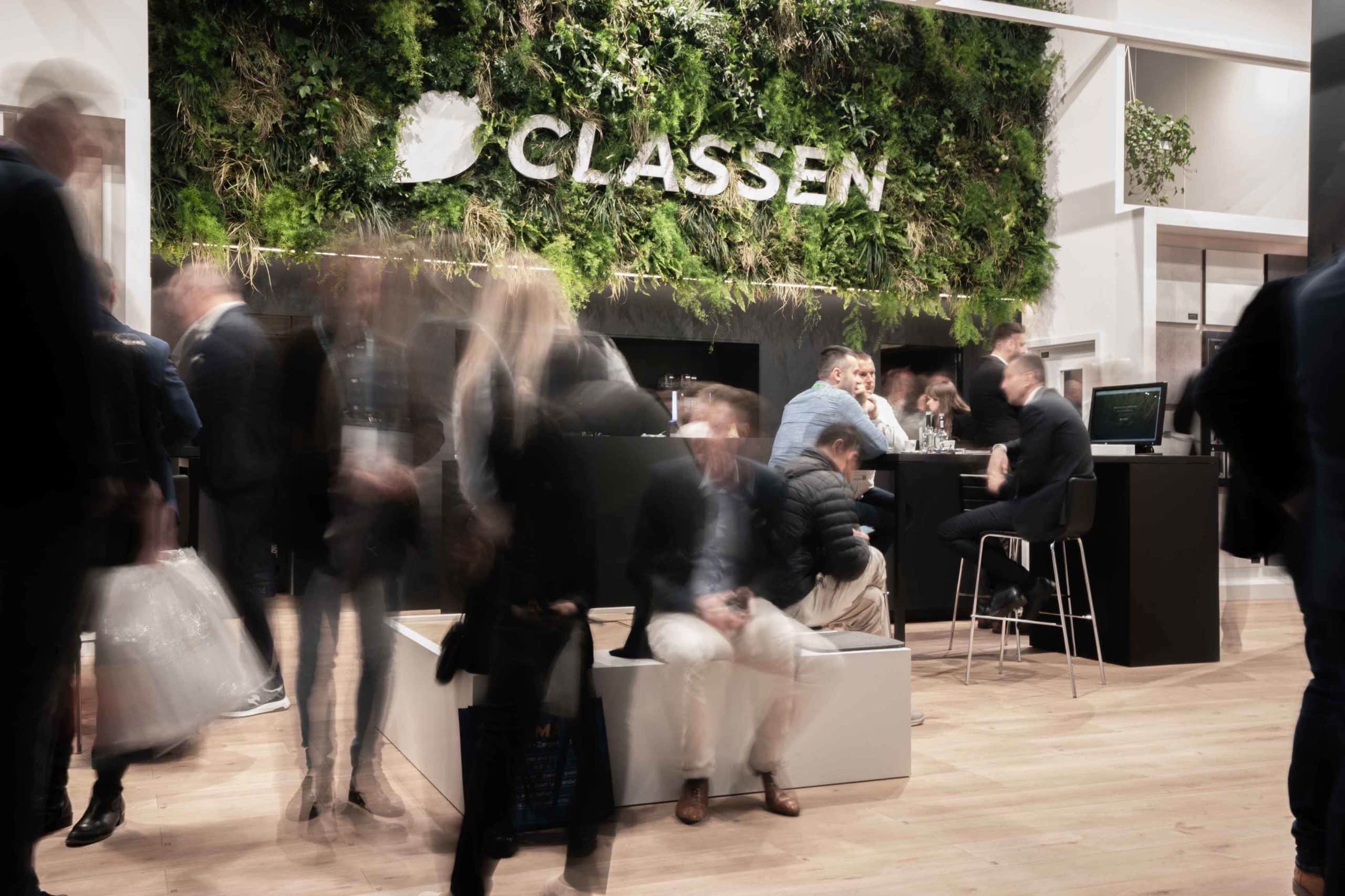 Besucherinnen und Besucher am CLASSEN Messestand mit auffälliger grüner Pflanzenwand und modernem Design. Menschen im Gespräch und Austausch über nachhaltige Bodenbeläge und Innovationen von CLASSEN.