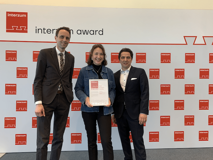 Drei Personen bei der Verleihung des interzum award: In der Mitte eine Frau mit Urkunde, flankiert von zwei Männern im Anzug. Im Hintergrund die offizielle interzum Award Wand. Symbol für Innovation und Auszeichnung in der Möbel- und Innenausbau-Branche.